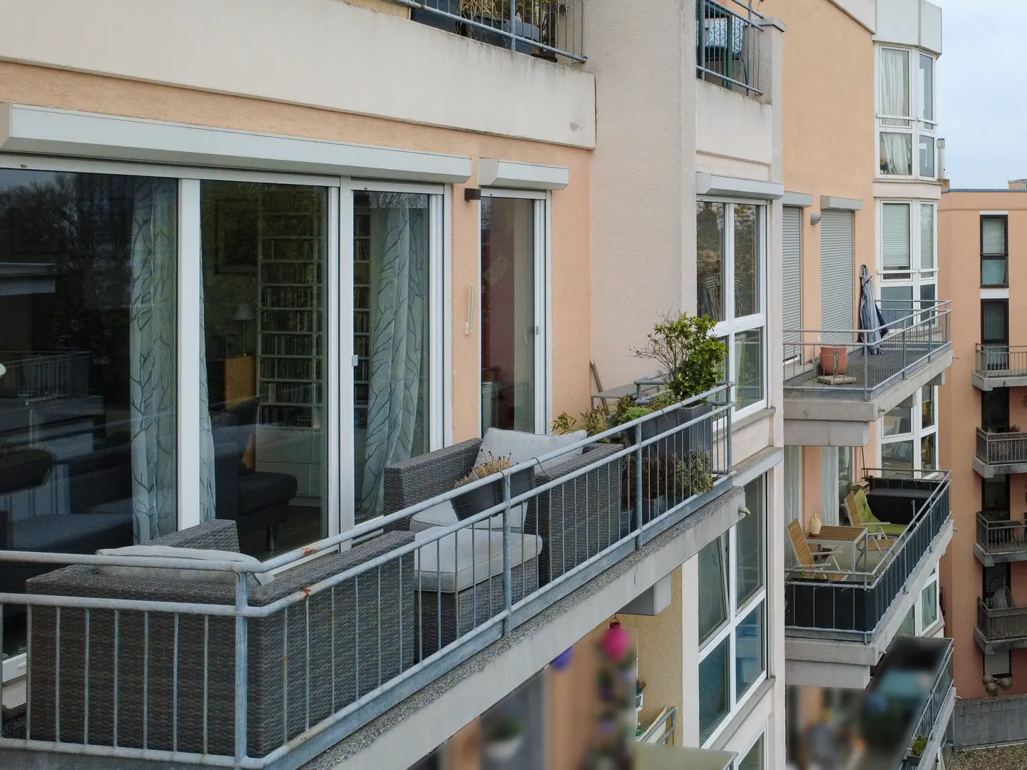 Apartment building exterior with balconies. Wicker patio furniture sits on the closest balcony. Other balconies have plants and furniture.