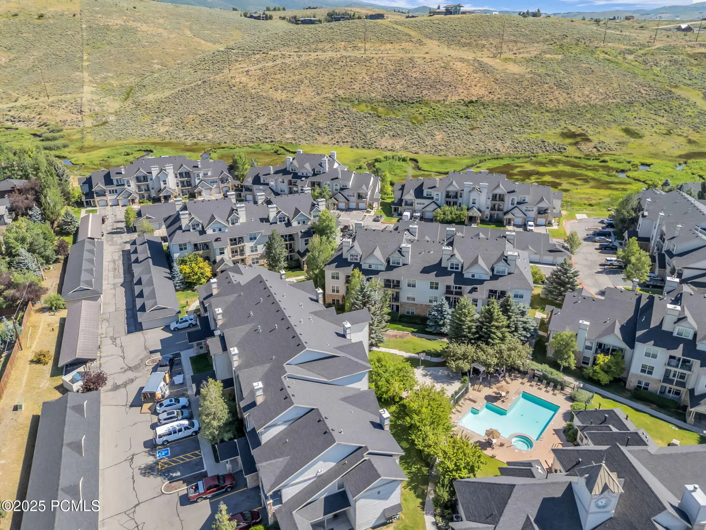 Aerial view of a condo complex with a pool, parking, and a grassy hill in the background.