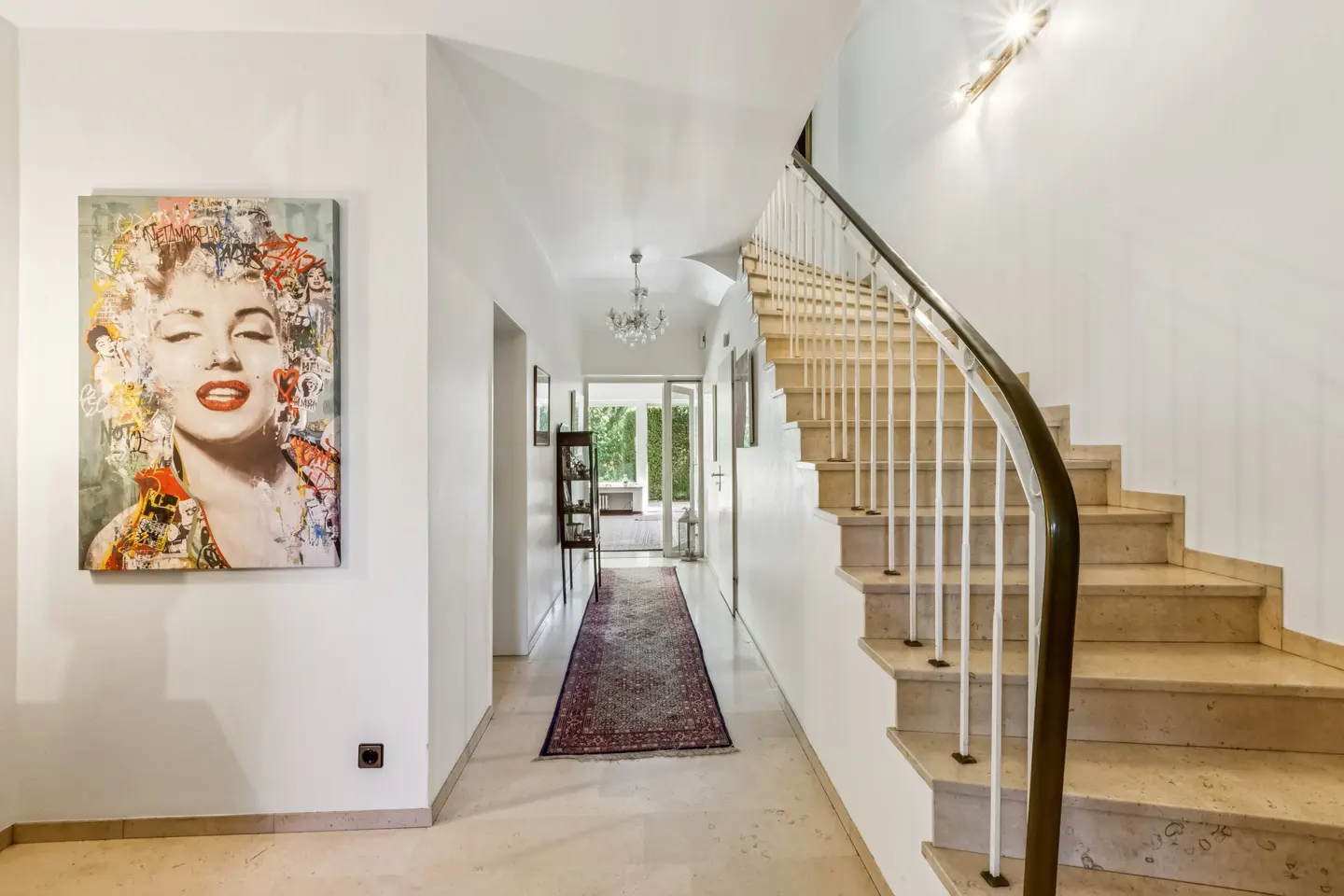 Bright hallway with marble floors, white walls, and a staircase. A Marilyn Monroe painting hangs on the left wall. A rug leads to a door at the end.