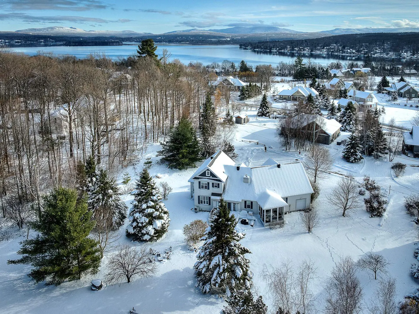 Aerial view of a snow-covered neighborhood with a white house, trees, and a lake in the background on a sunny day.