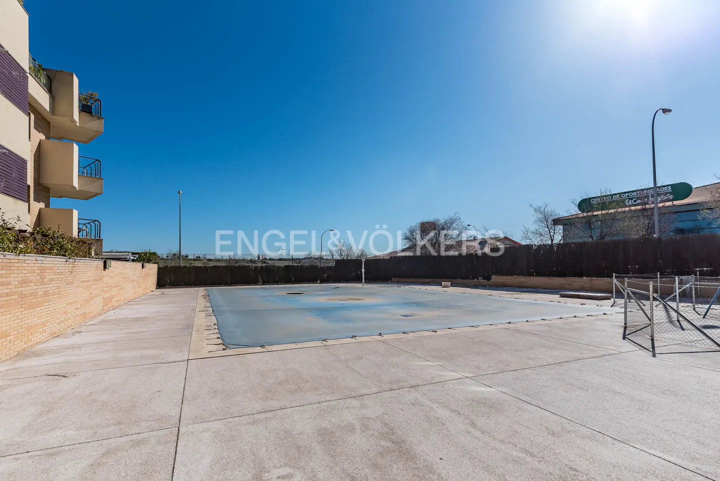 Outdoor pool covered with a blue tarp, surrounded by a concrete patio and a brick wall, under a clear blue sky.