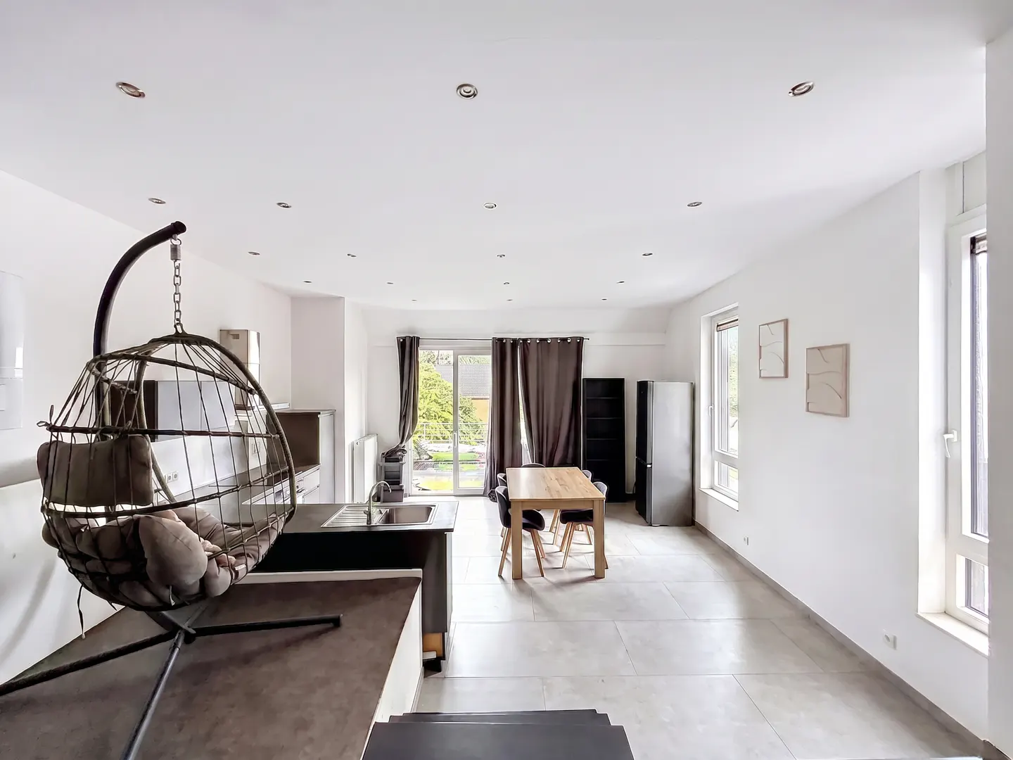 Bright, open-plan living space with white walls, tiled floor, and a hanging chair. Dining table, kitchen area, and garden view through sliding doors.