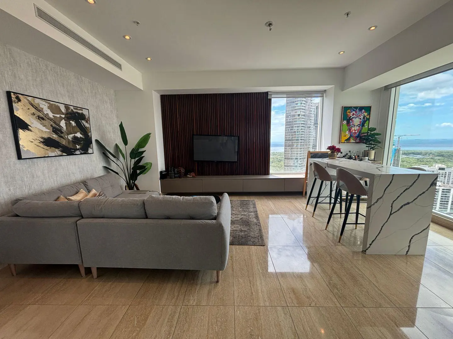 A modern living room with a gray sectional sofa, a TV on a wood-paneled wall, and a marble island with bar stools. A large window shows a city view.
