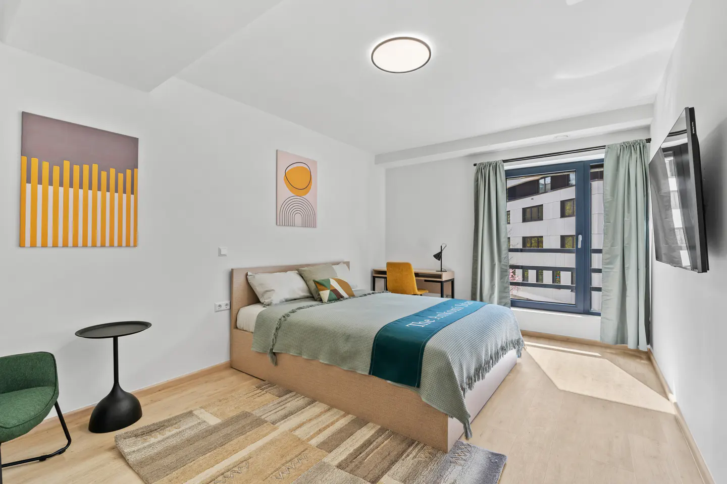 Bright bedroom with a bed, desk, and chair. Artwork hangs on the white walls. A window with curtains overlooks a building.