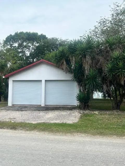 A white two-car garage with a red roof, set back from the road with trees.