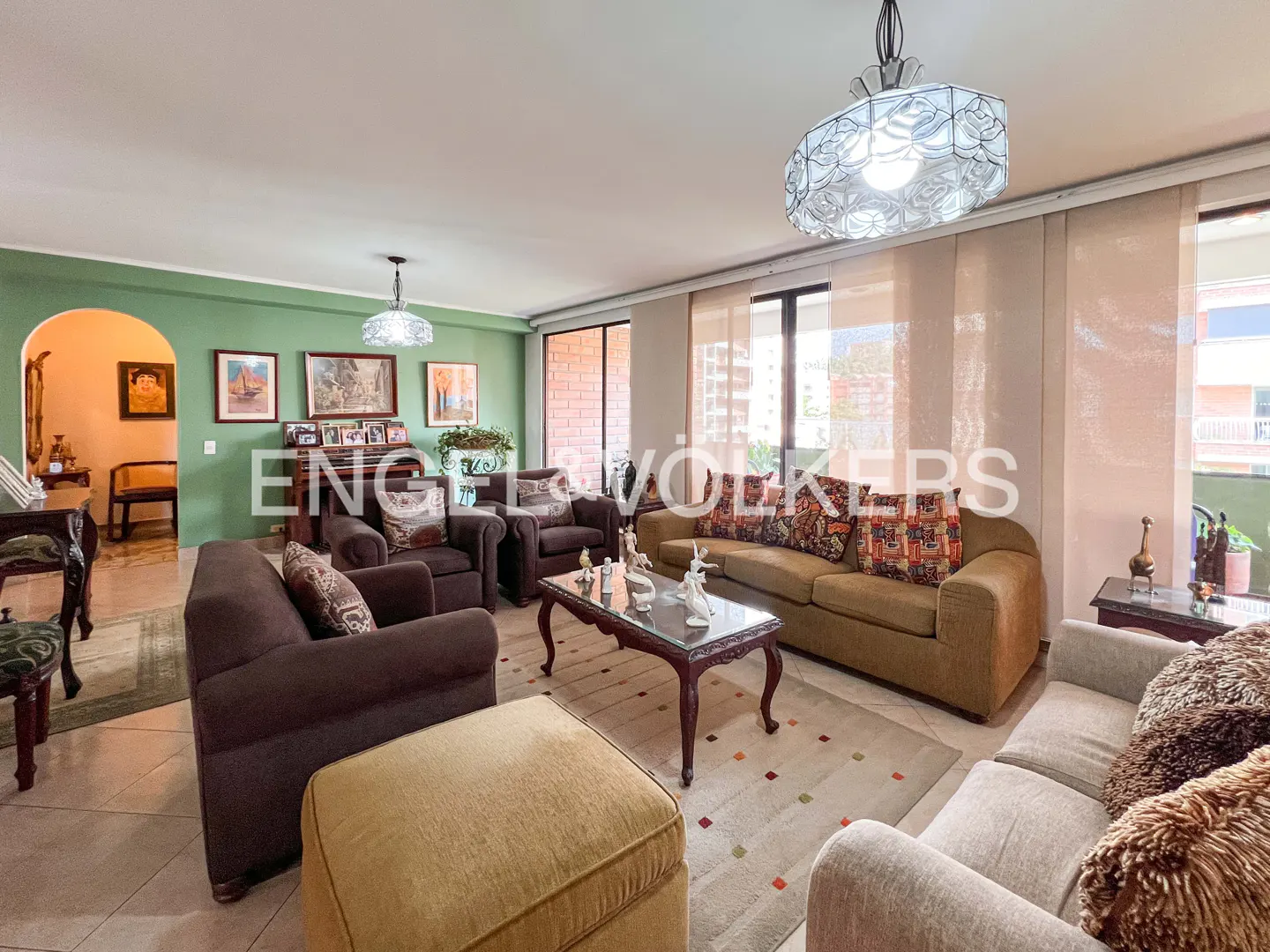 A spacious living room with brown sofas, a coffee table, and a light-colored rug. The walls are green, and there are windows with sheer curtains.