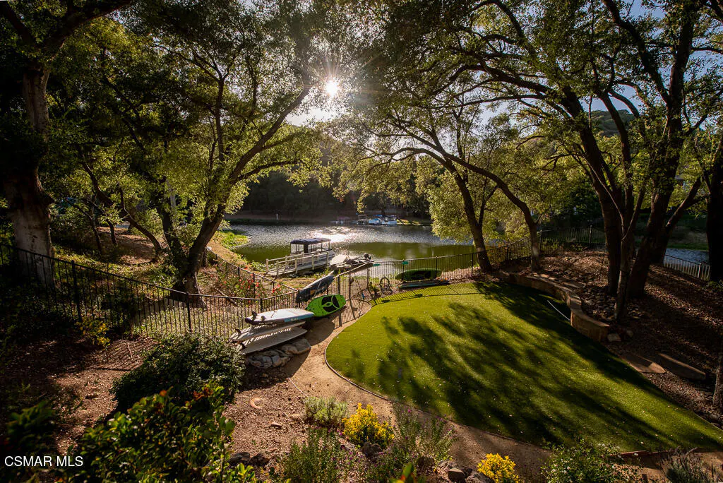 Scenic view of a lake with a dock, boats, and kayaks, framed by trees and a green lawn. Sunlight filters through the leaves.