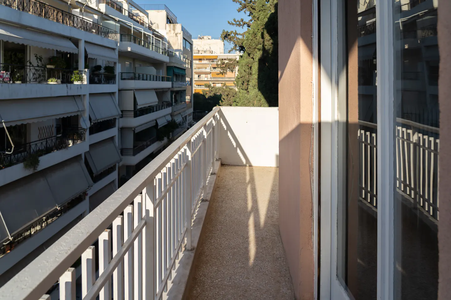 A narrow apartment balcony with a white railing, overlooking a street lined with multi-story residential buildings.