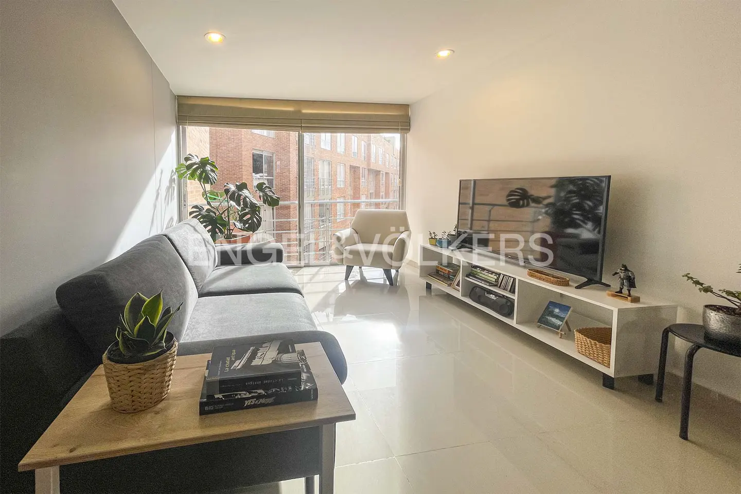 Bright living room with gray sofa, beige armchair, and TV on a white console. Large window with view of brick building.