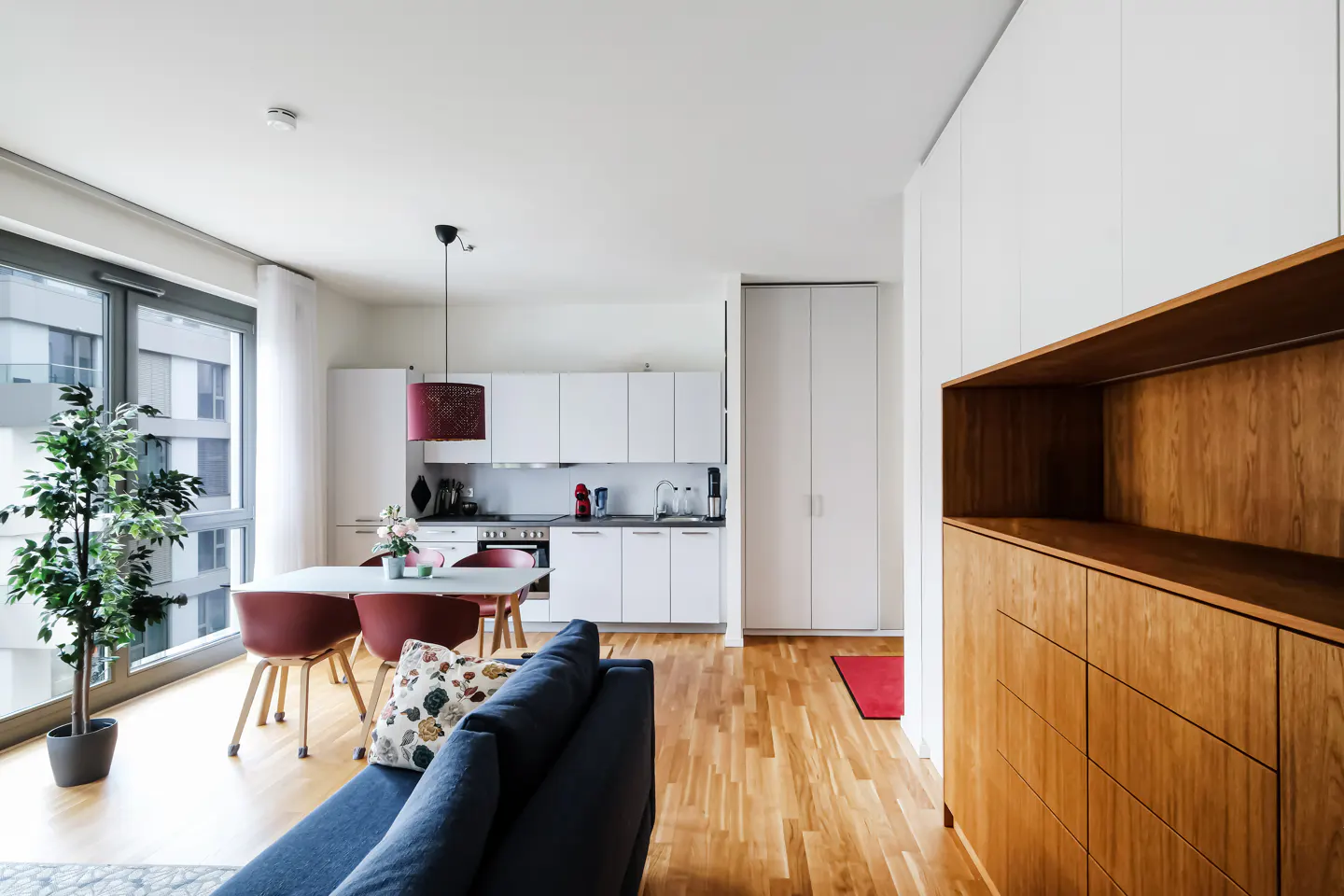 Bright apartment interior with hardwood floors, white kitchen cabinets, dining table with red chairs, and a blue sofa.