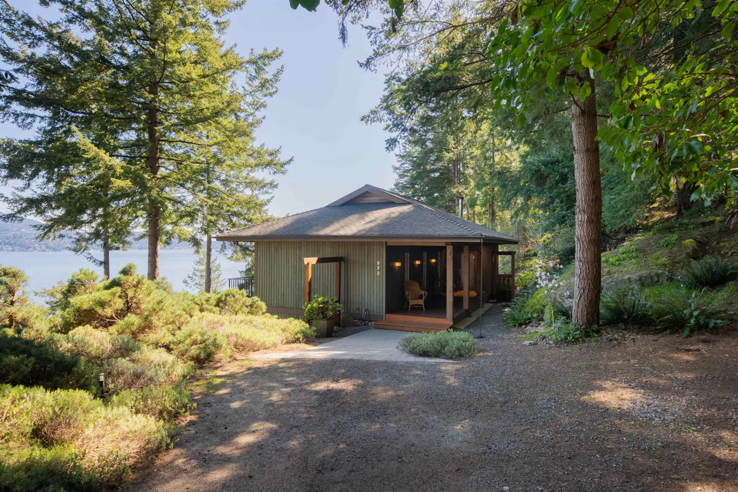 Exterior view of a one-story, light green house with a covered porch, surrounded by trees and greenery.