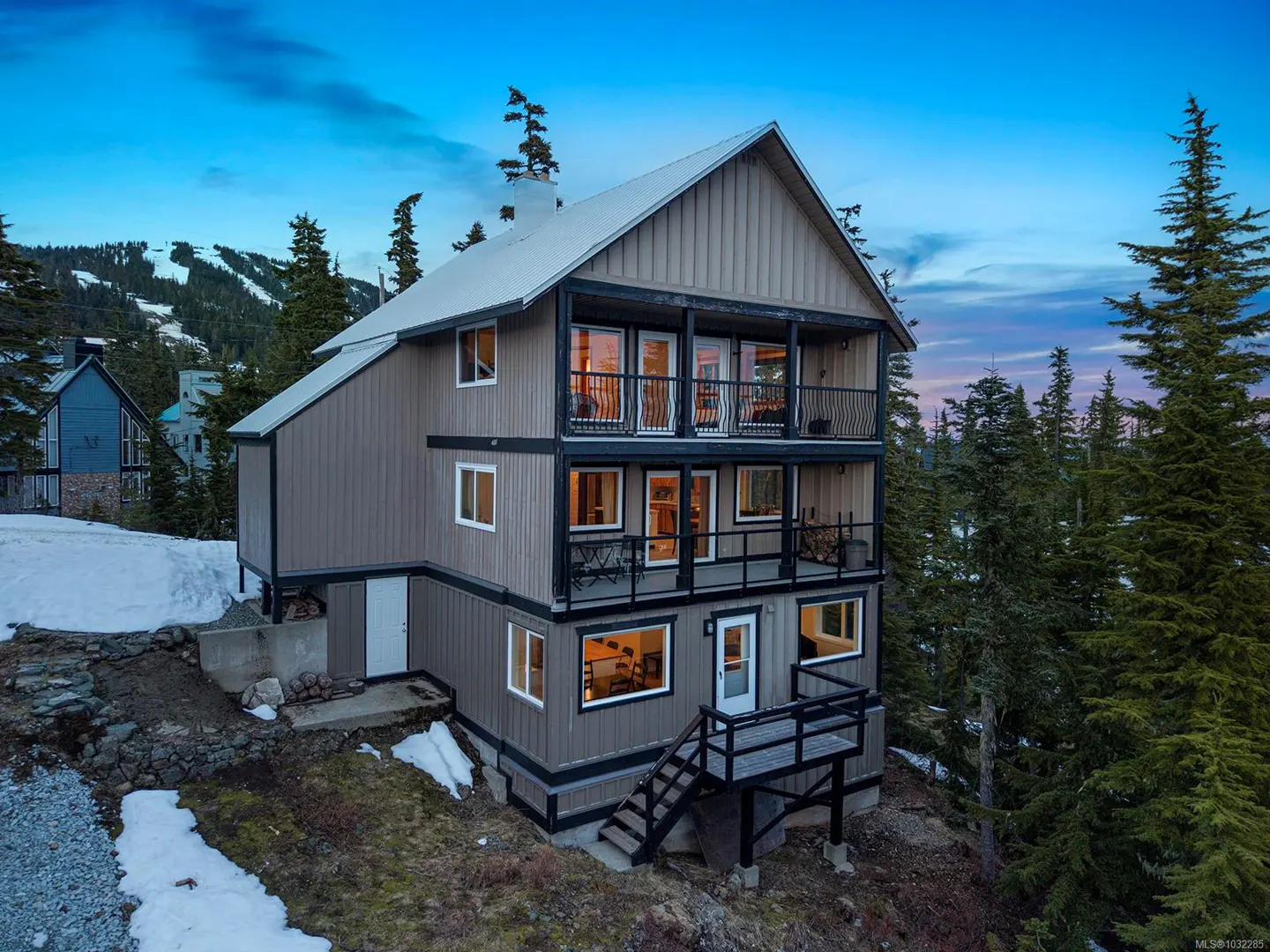Three-story brown house with black trim and balconies, nestled in a snowy, wooded area under a blue sky.