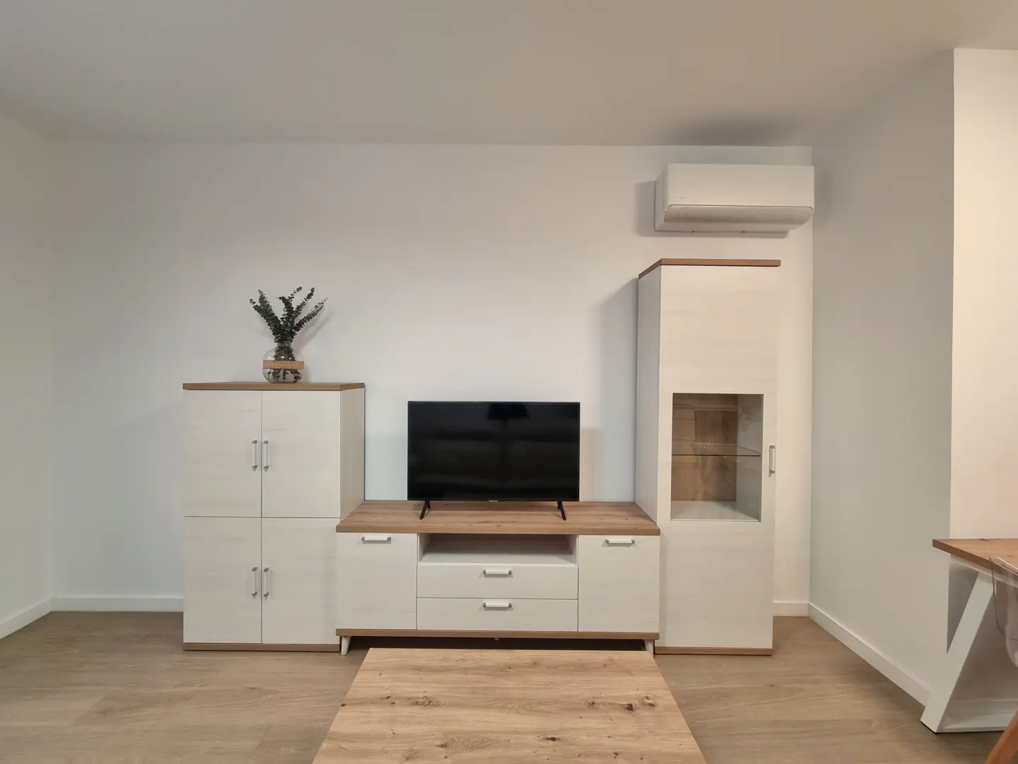 Living room with white walls, light wood floors, and matching cabinets. A TV sits on a low cabinet, and a coffee table is in front.