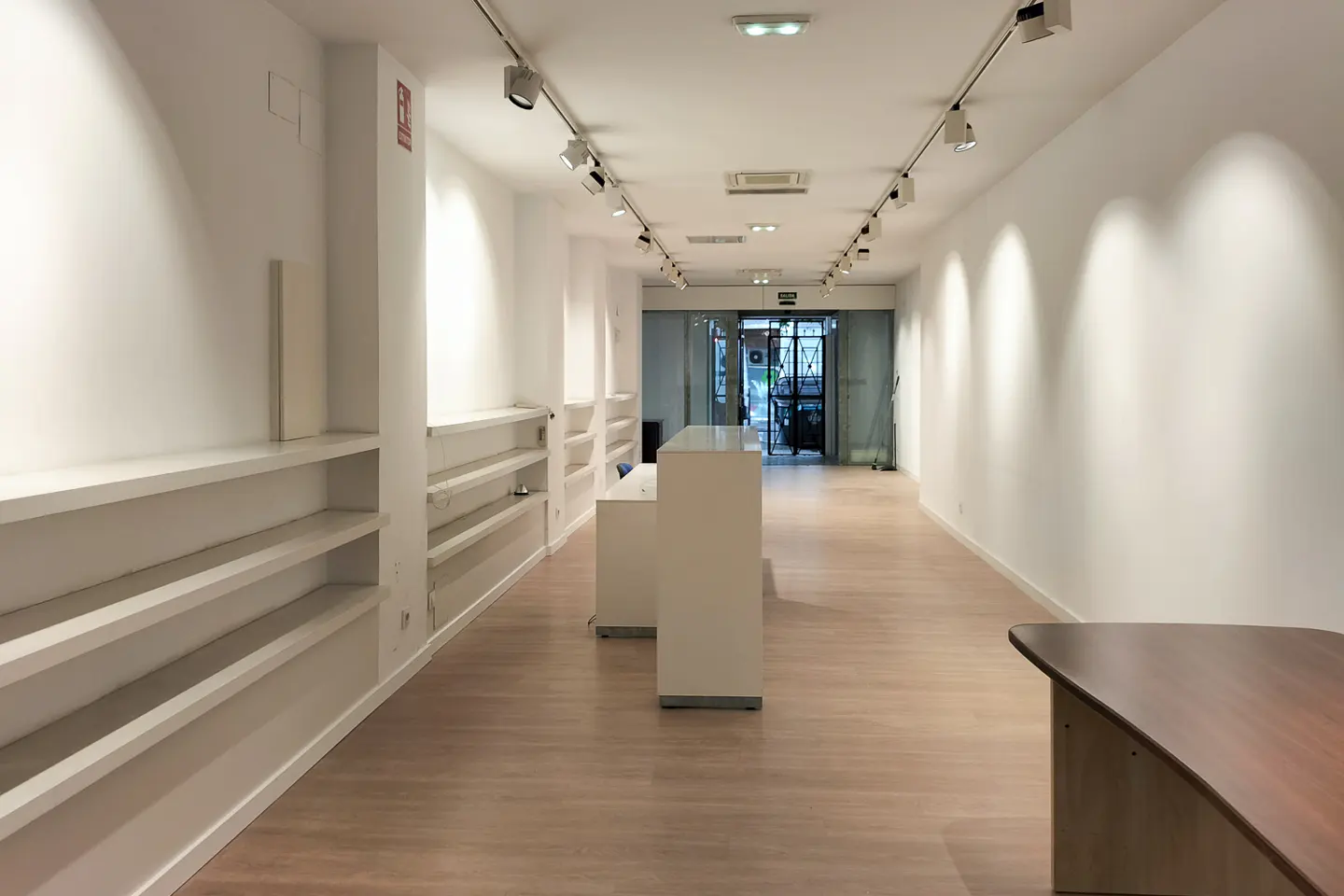 Bright, empty retail space with white walls, shelves, and light wood floors. Track lighting runs along the ceiling. A white counter sits in the middle. Glass doors at the back.