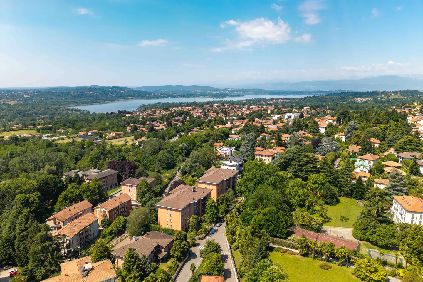 Aerial view of a lush Italian town with orange-roofed buildings, dense trees, and a large lake under a clear blue sky.