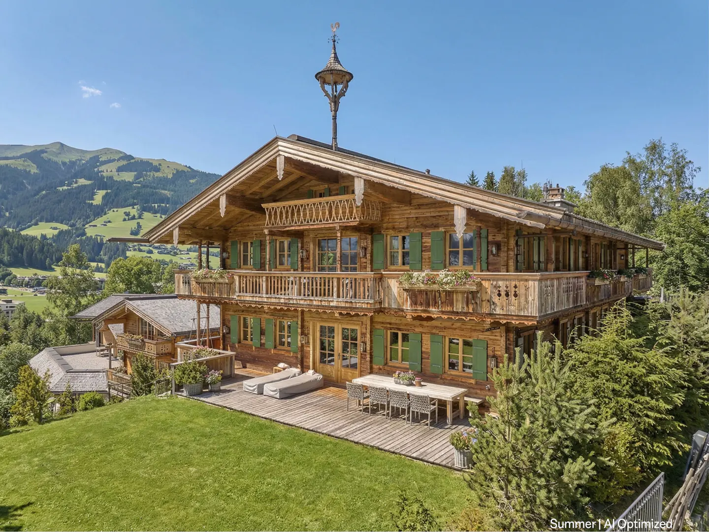 A large, traditional wooden chalet with green shutters and a balcony, set against a scenic mountain landscape.