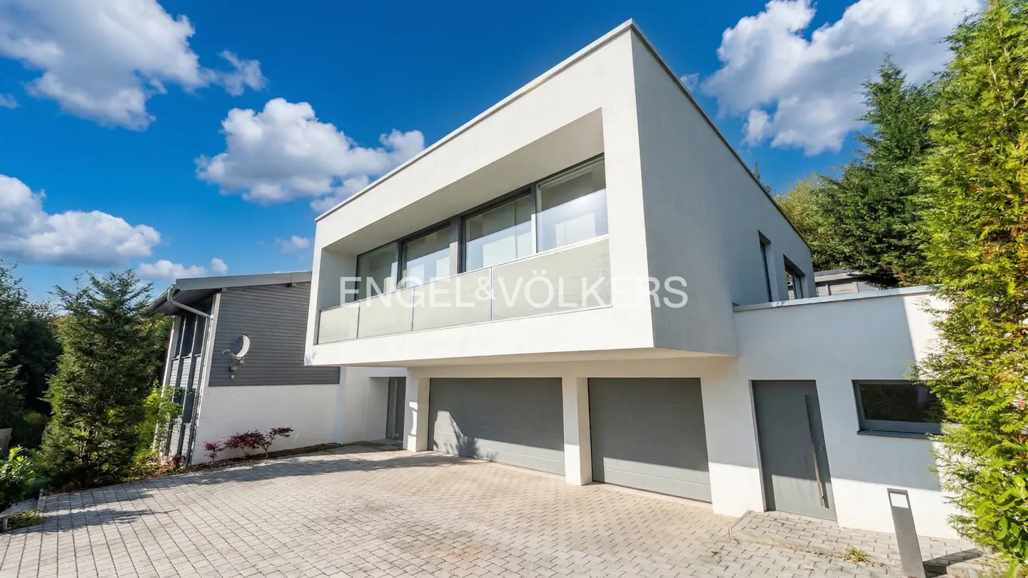 Modern white house with gray garage doors and a glass balcony under a blue sky with clouds.