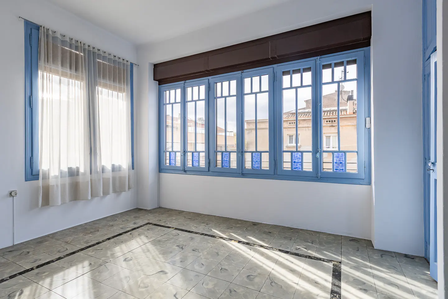 Bright room with white walls, patterned tile floors, and large blue-framed windows with sheer curtains and city views.