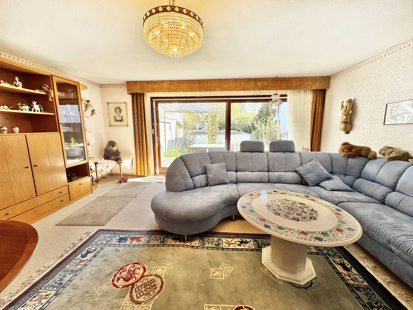 Living room with a blue sectional sofa, a round marble table, and a large patterned rug. A chandelier hangs from the ceiling. Sliding glass doors lead to a garden.