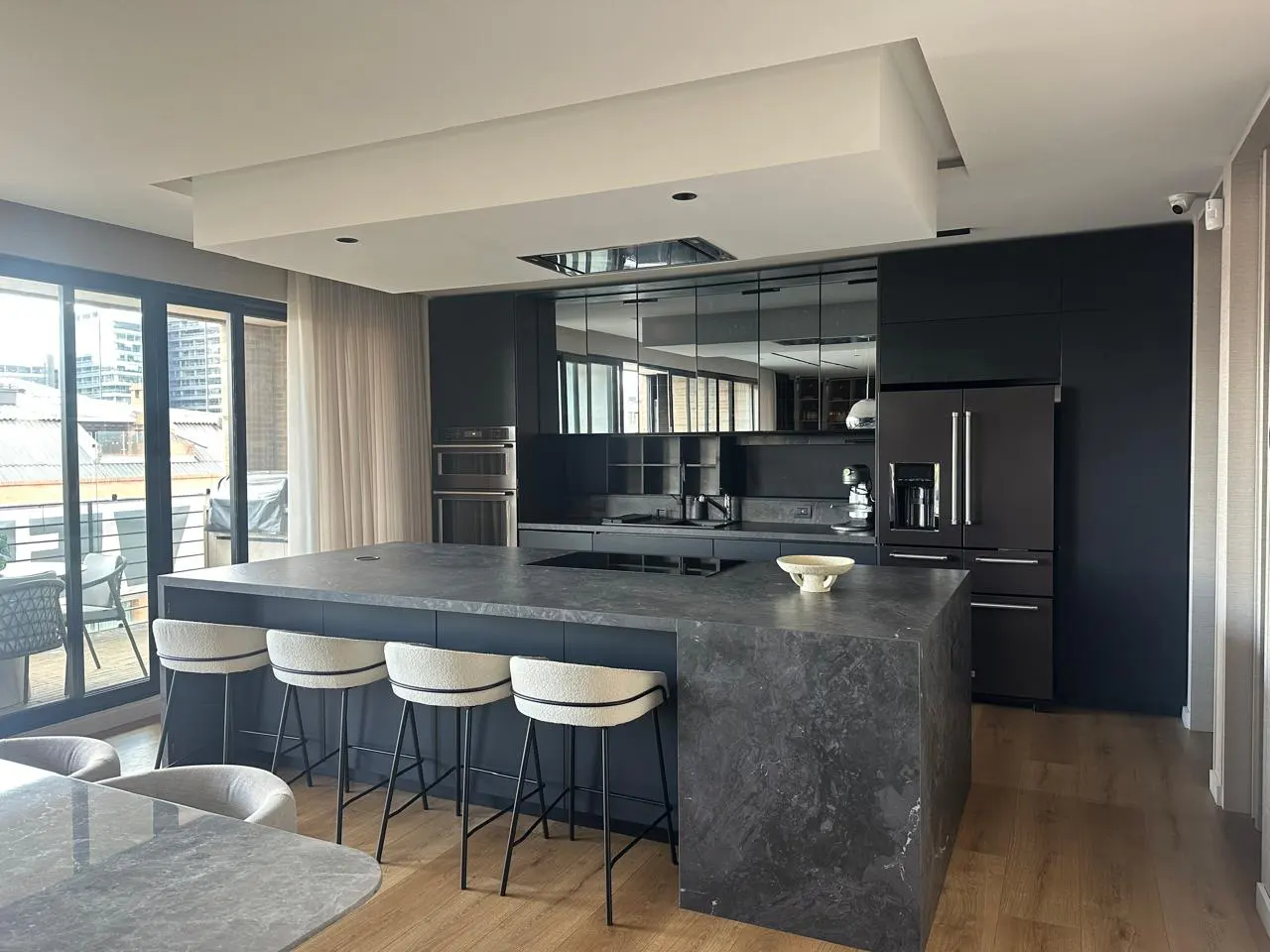 A modern kitchen with dark cabinets, a large marble island with four stools, and a view of the city through a window.