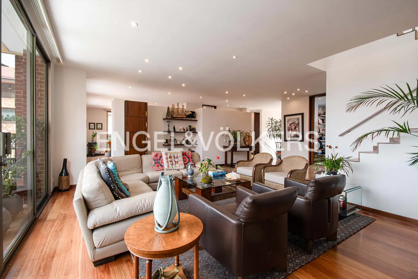 Bright living room with a beige sectional sofa, brown leather chairs, and a glass coffee table on a patterned rug. Hardwood floors and large windows.