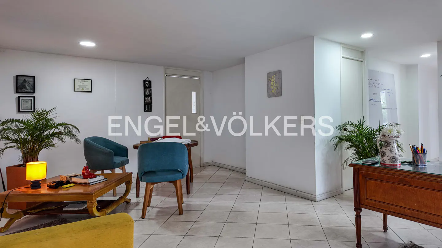 Bright office space with teal chairs, wooden tables, and potted plants against white walls and tiled floors.