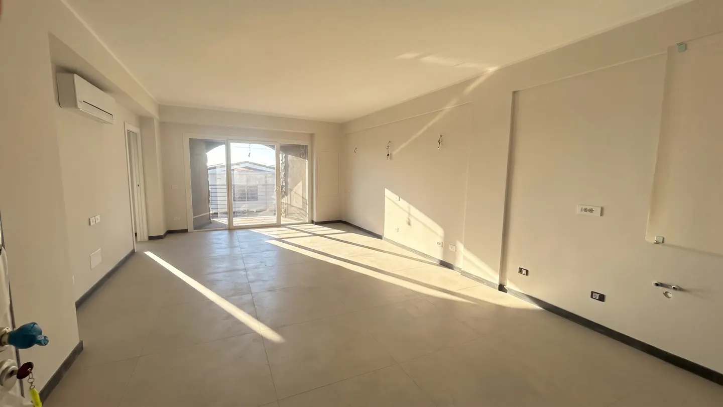Bright, empty room with white walls and tile floor. Sunlight streams in from sliding glass doors to a balcony.