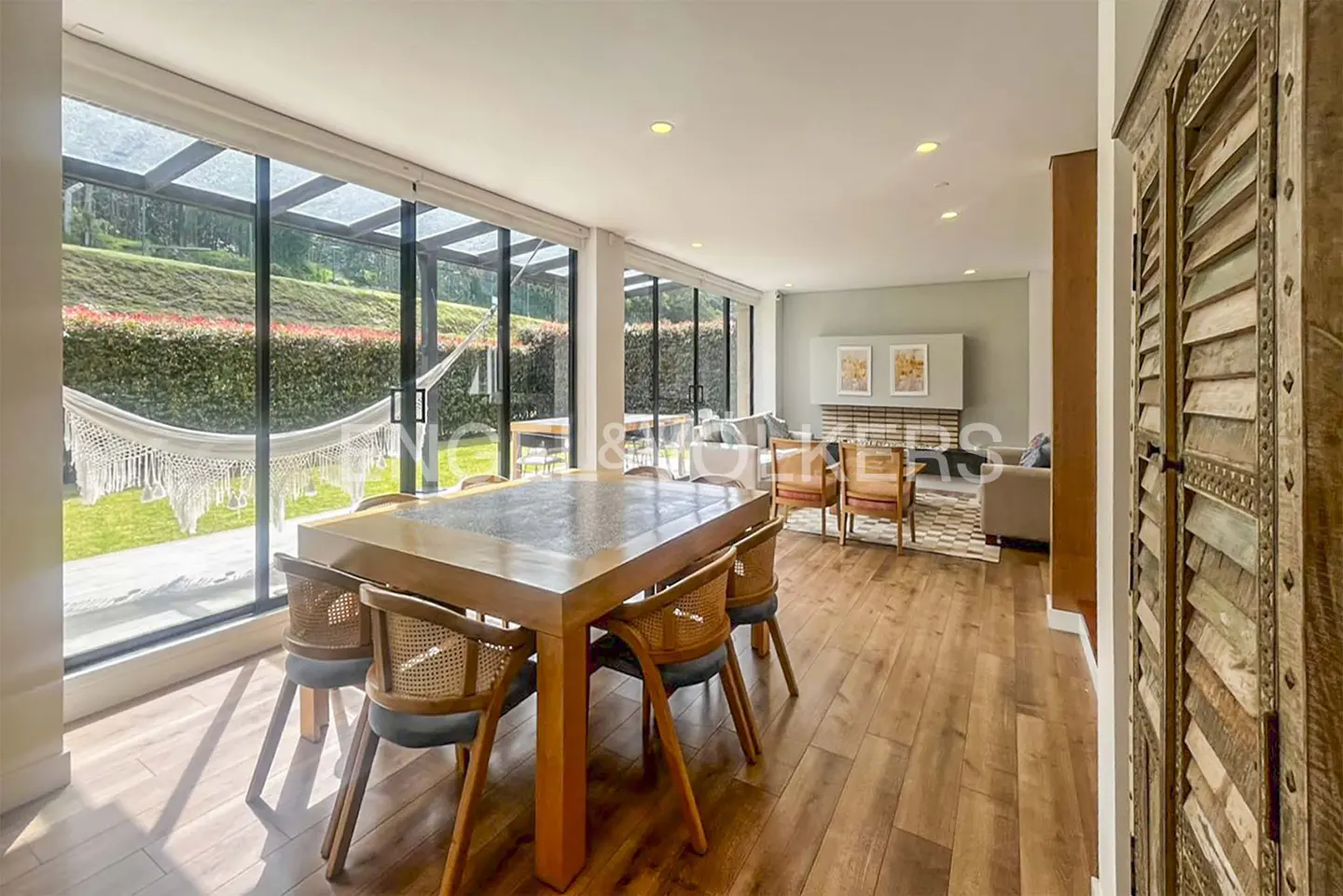 Bright, open dining and living area with wood floors. A large table sits near windows overlooking a green lawn with a hammock.