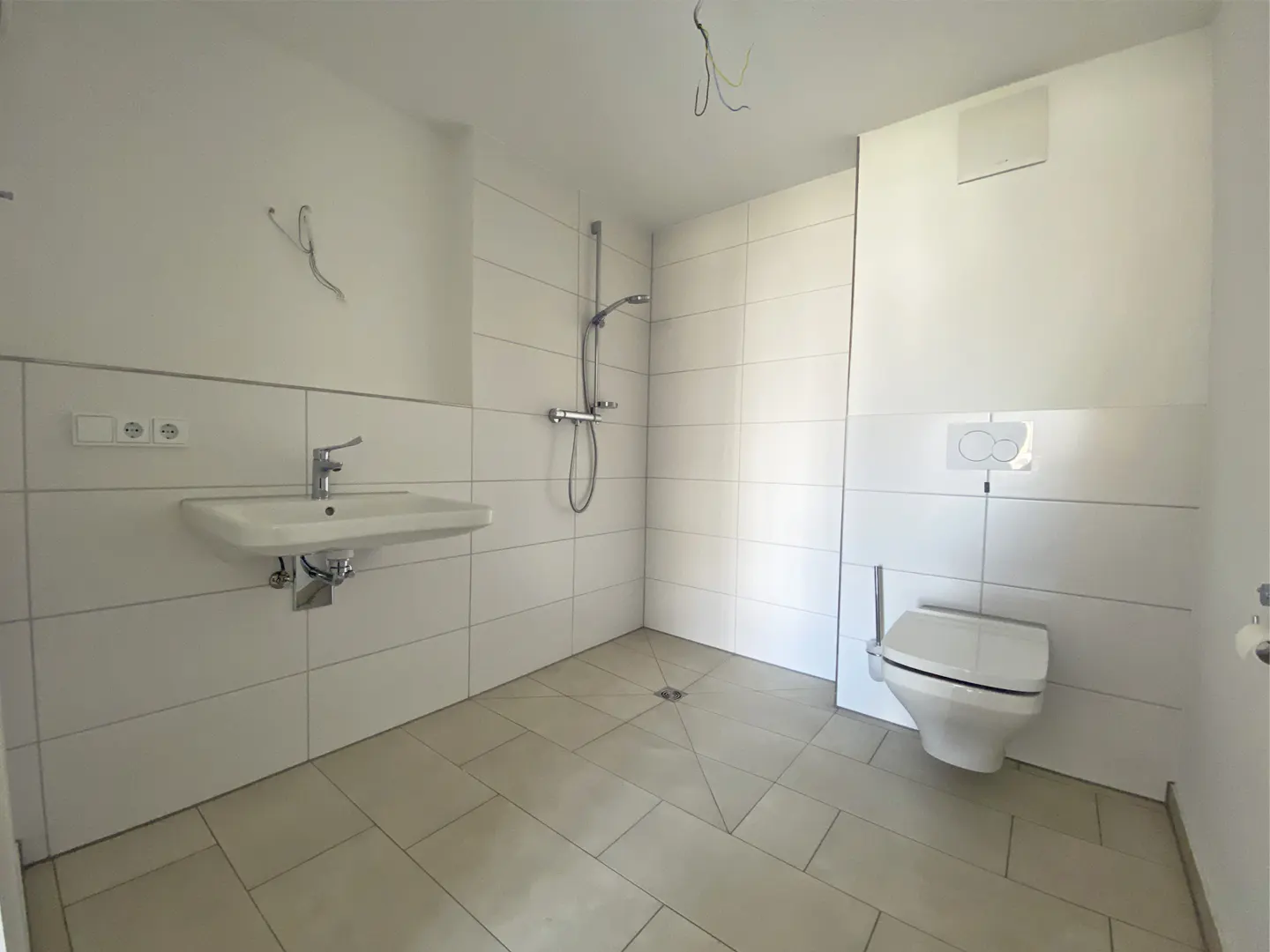 Bright bathroom with white tiles, sink, shower, and toilet. Beige floor tiles and exposed wiring in the ceiling.