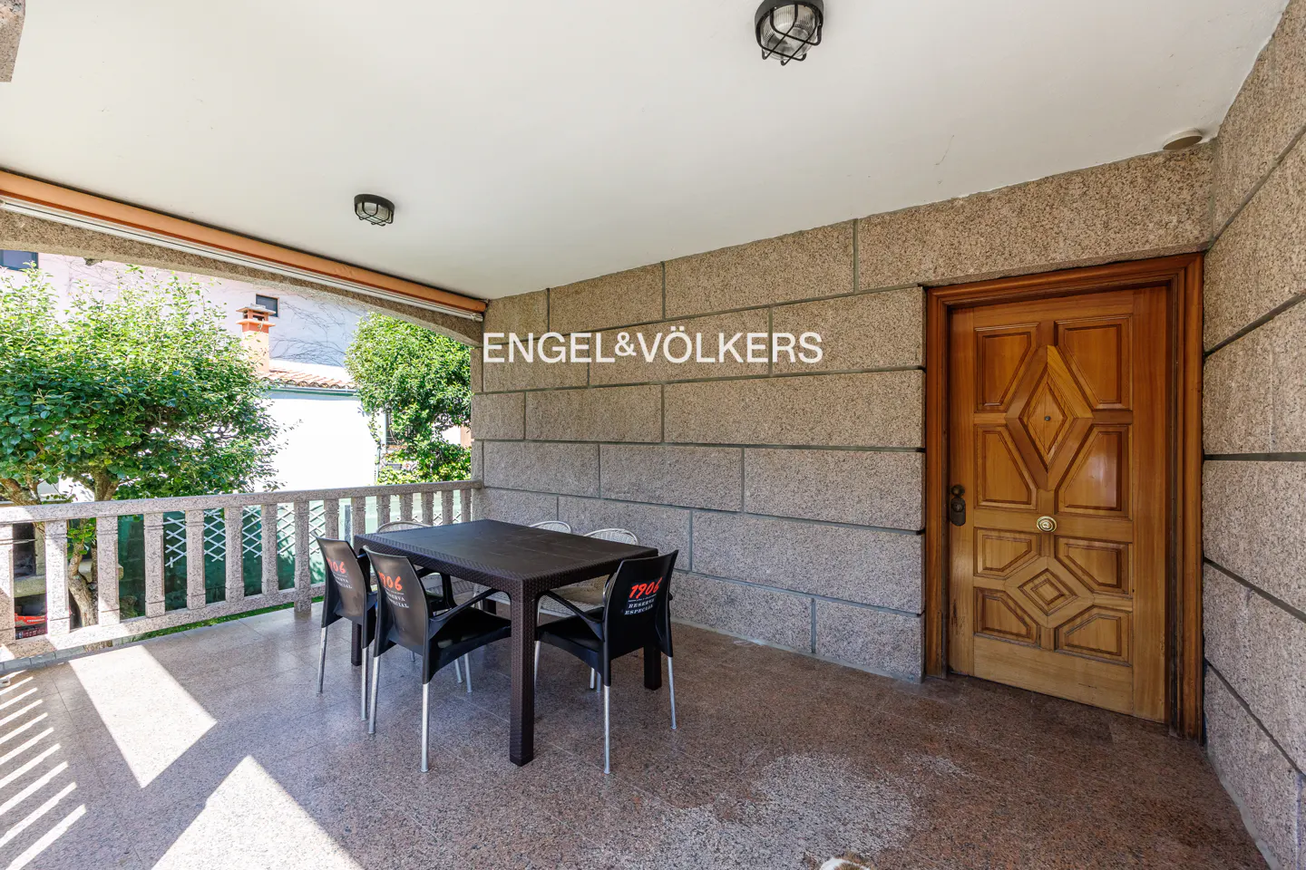 Covered stone patio with a dining table, four black chairs, and a wooden door, overlooking a garden with a stone railing.