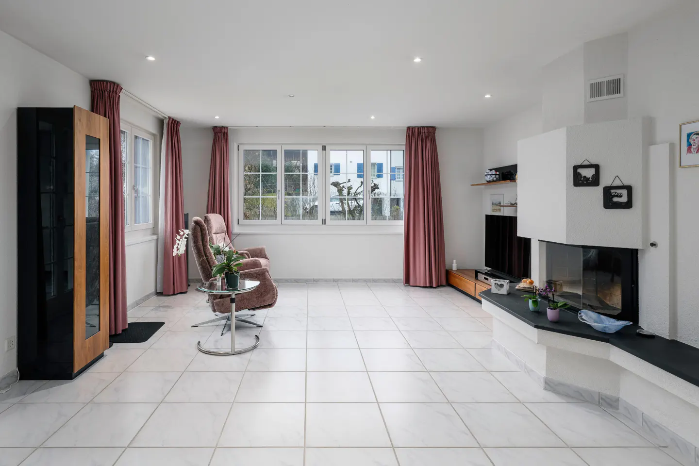 Bright living room with white tile floor, pink curtains, and a modern fireplace. A brown armchair sits near a window.