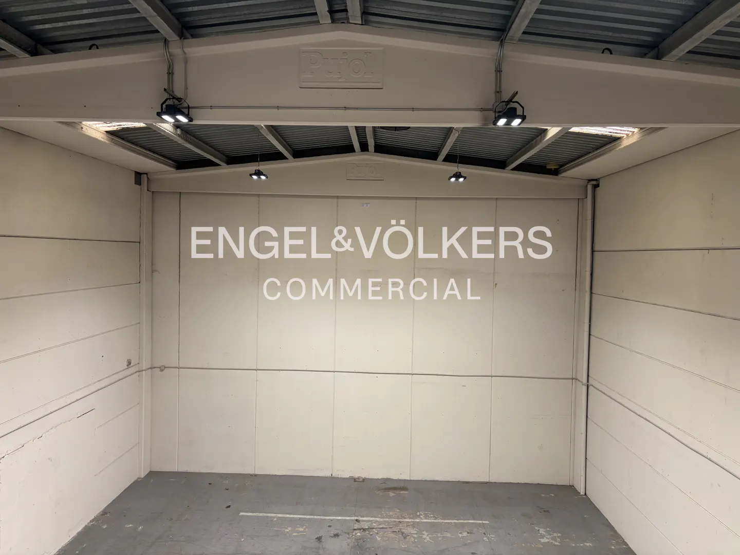 Empty commercial space with "Engel & Völkers Commercial" in white on the back wall. Gray concrete floor and metal ceiling with lights.