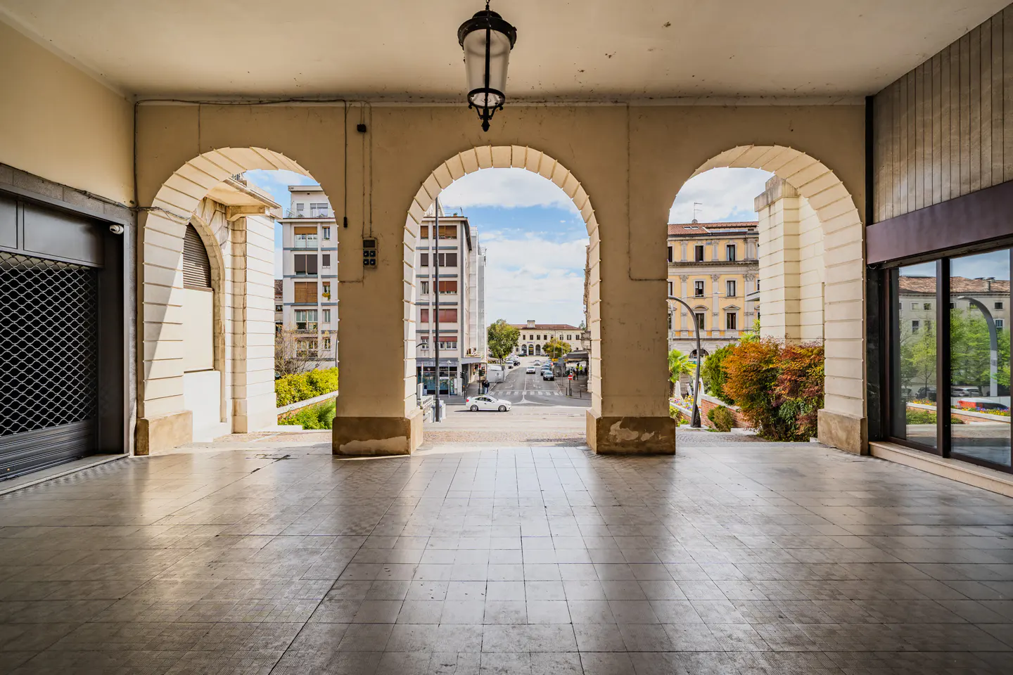 View through three arches of a building, showing a street with cars and buildings in the distance on a sunny day.