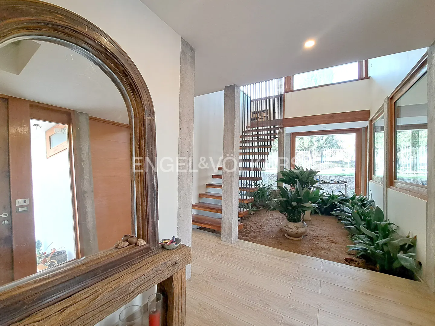 Modern foyer with a rustic wooden mirror, floating staircase, and an indoor garden with potted plants on a dirt floor.
