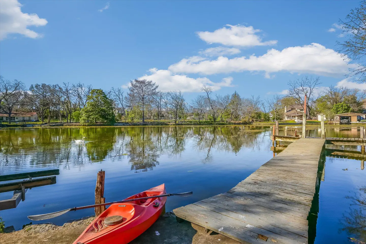 Scenic lake view with a red kayak, wooden dock, and a swan swimming in the water under a blue sky with clouds.