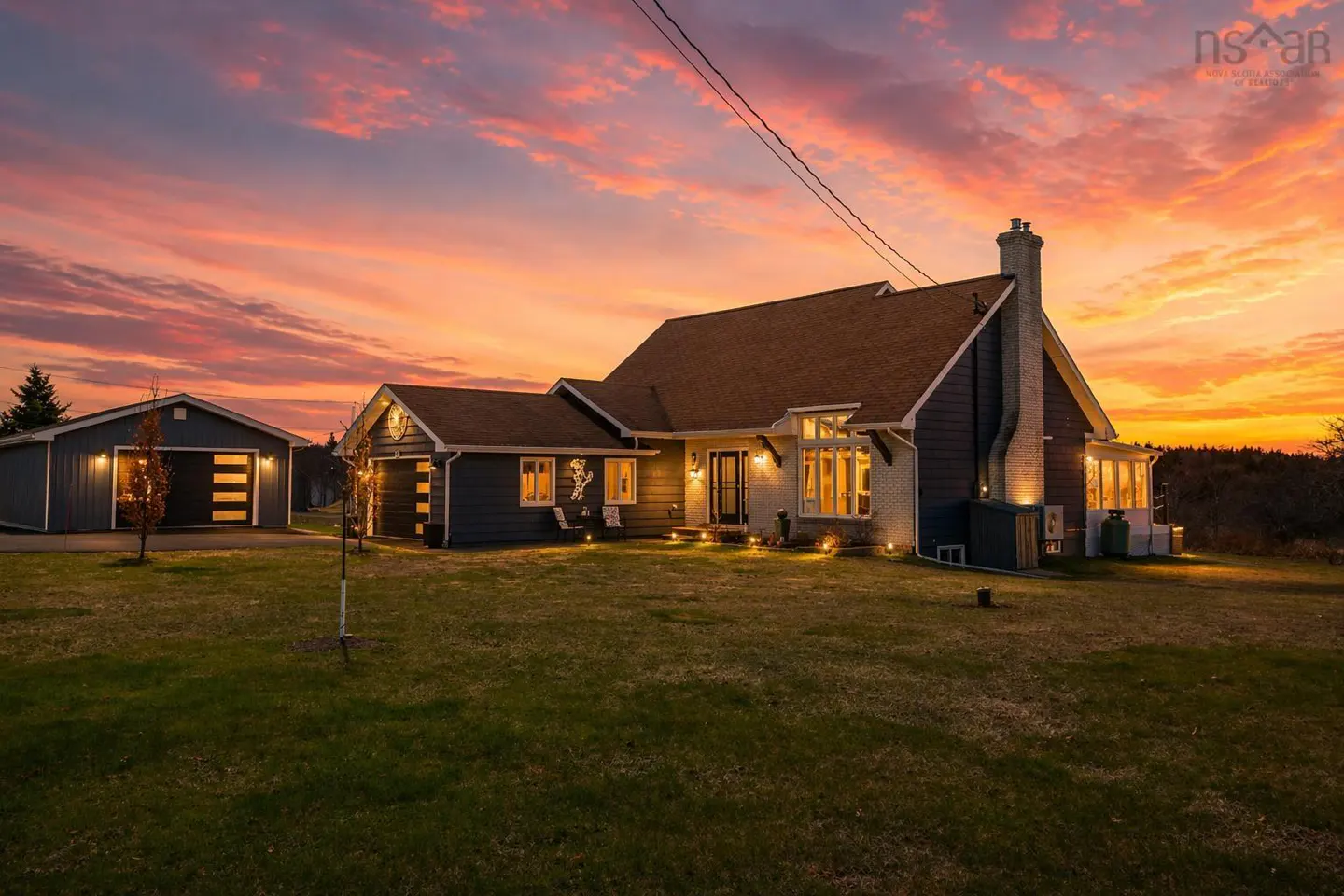 A single-family home with a detached garage at sunset. The sky is orange and pink. The house is dark blue with a brown roof.