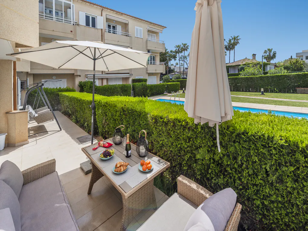 Patio with wicker furniture, table set with food, and a view of a pool and manicured hedges on a sunny day.