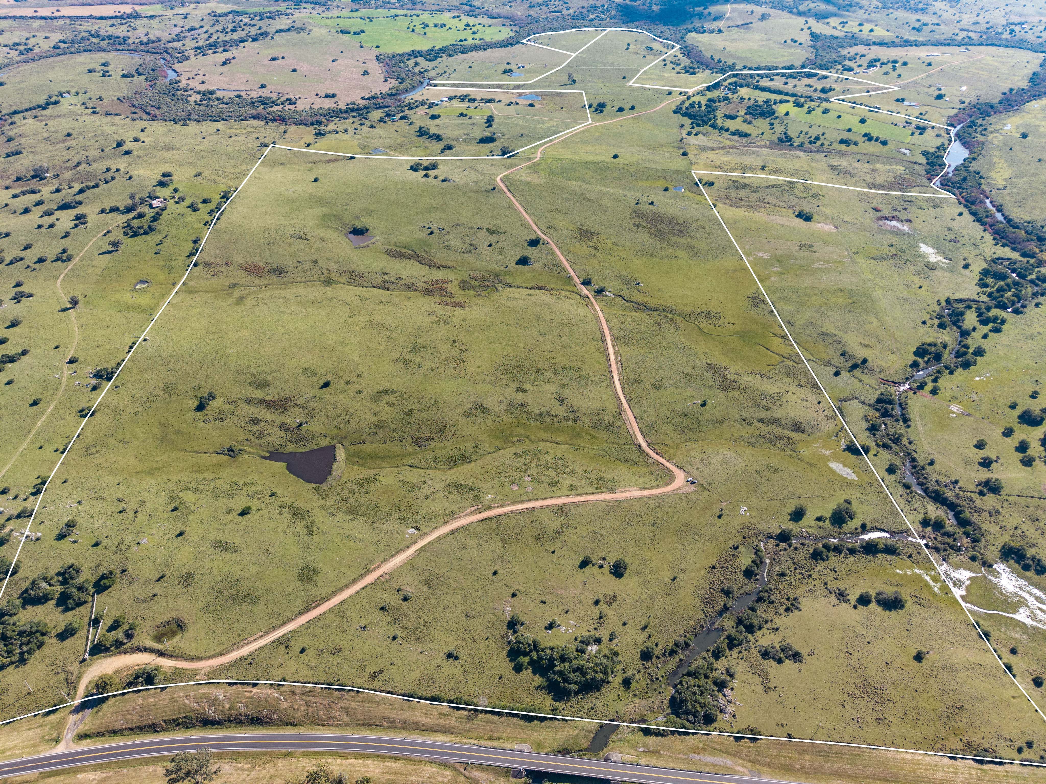Aerial view of a large, green rural property outlined in white, featuring a dirt road, small ponds, and a nearby highway.