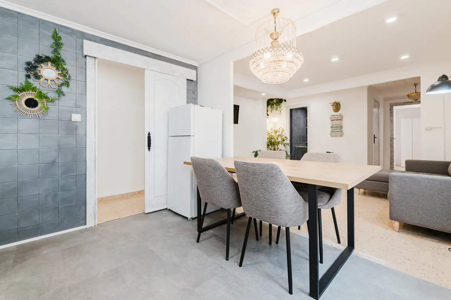 A bright dining area with a light wood table, gray chairs, and a crystal chandelier. A gray tiled wall with decorative mirrors adds texture.