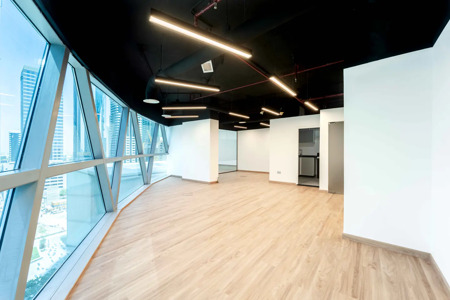 Bright, empty office space with wood floors, white walls, and a black ceiling with linear lights. Large windows offer a city view.