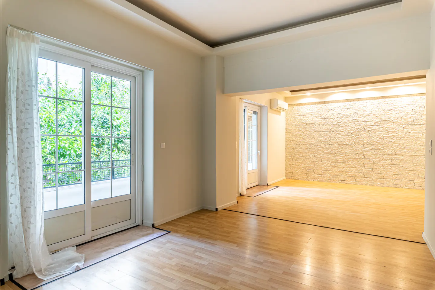 Bright, empty room with wood floors, white walls, and a textured stone accent wall. A glass door leads to a balcony with green foliage visible.