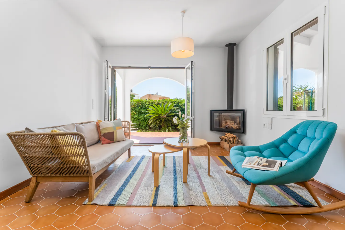 Bright living room with a wicker sofa, blue rocking chair, fireplace, and open doors to a lush garden.