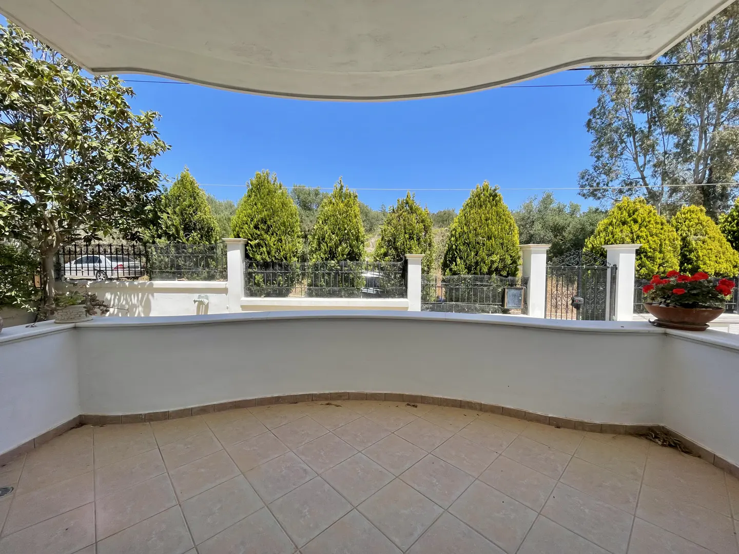 View from a tiled balcony with a white wall and black iron fence. Green trees and a clear blue sky are visible beyond the fence.