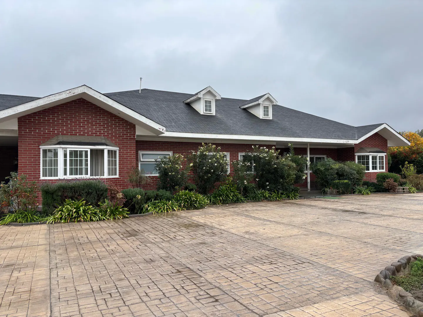 Red brick building with a dark gray roof and white trim, dormer windows, and a brick-paved driveway.