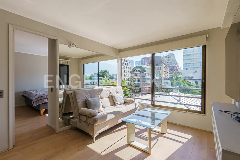 Bright apartment interior with a beige sofa, glass table, and city view through large windows. Bedroom visible through doorway.