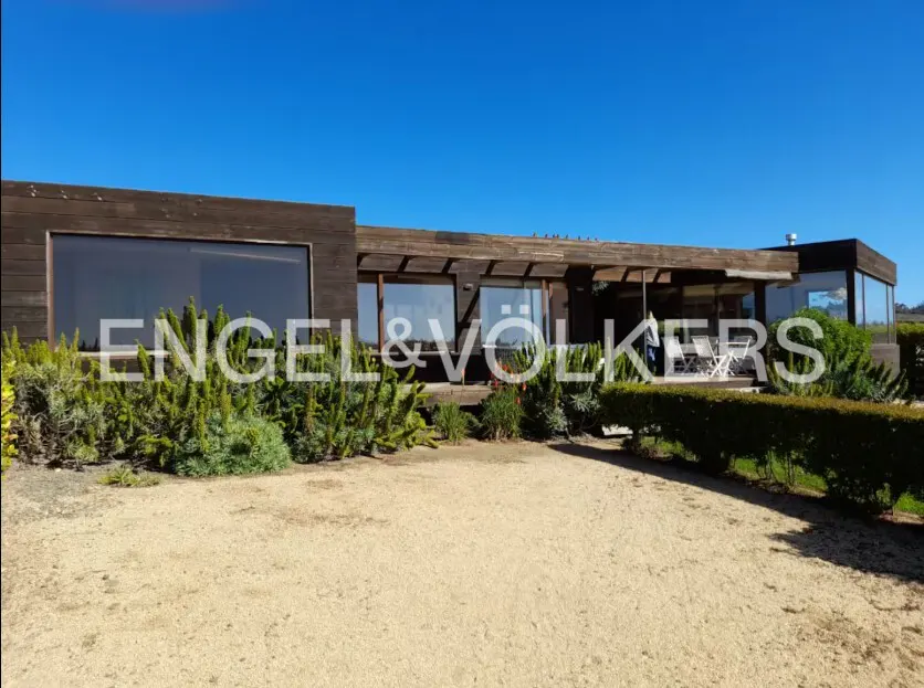 Exterior view of a modern, single-story brown house with large windows, green bushes, and a gravel yard under a clear blue sky.