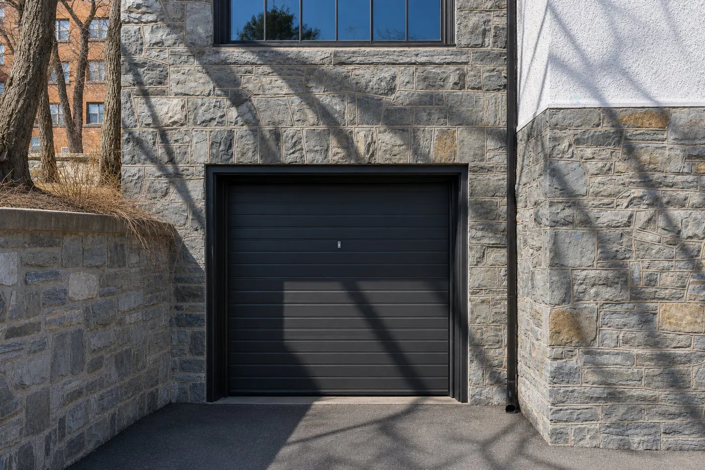 A gray, horizontal-paneled garage door is set in a stone wall with a window above.