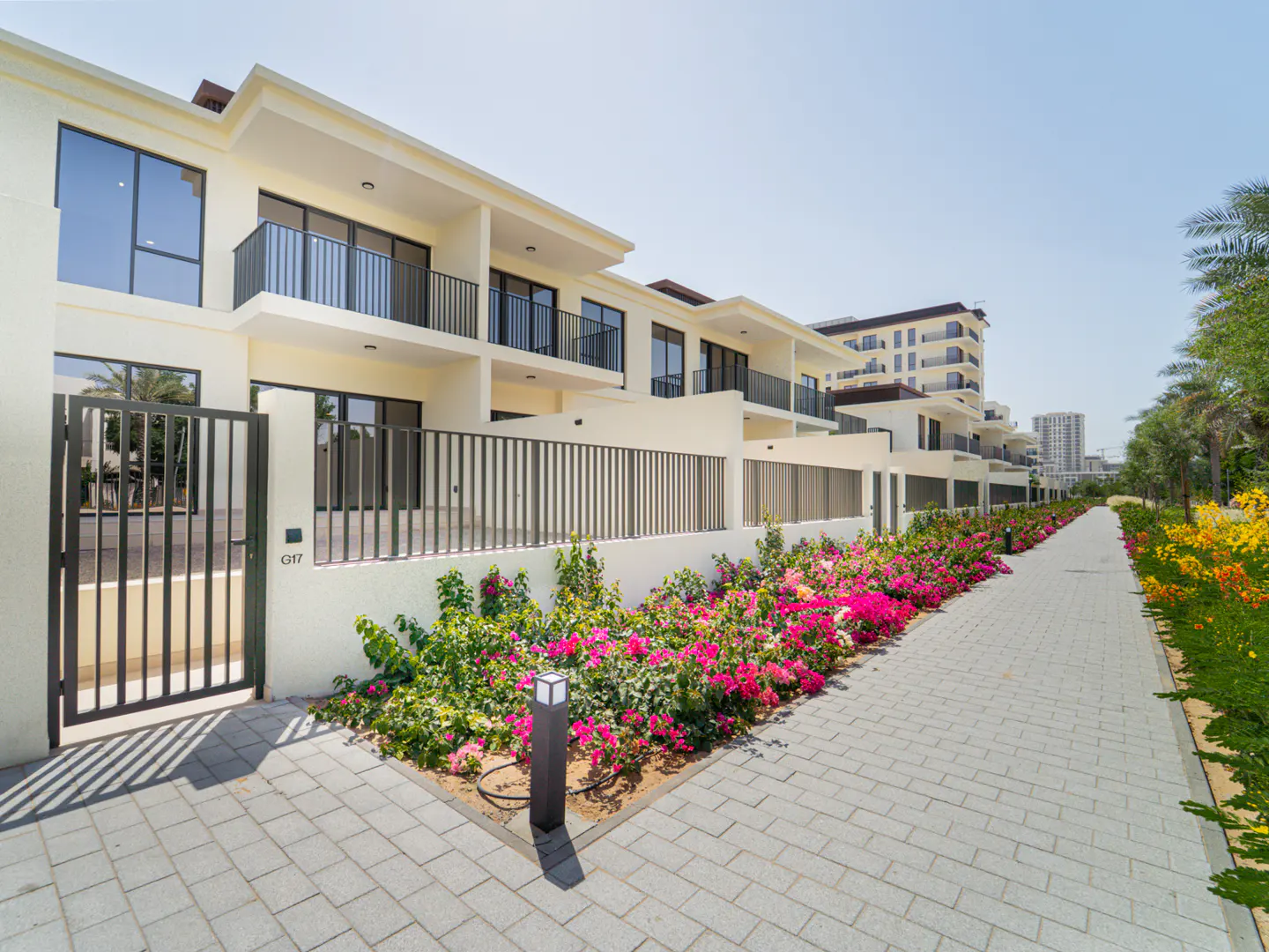 Row of modern, two-story townhouses with black railings and large windows. A brick walkway is lined with colorful flowers and greenery.