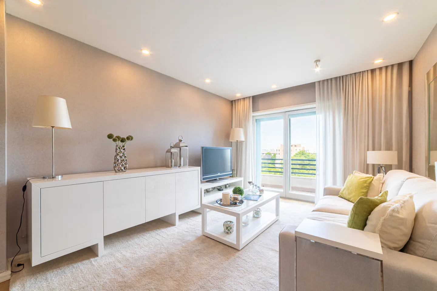 Bright living room with beige walls, white furniture, and a sliding glass door to a balcony. A cream-colored sofa has green pillows.