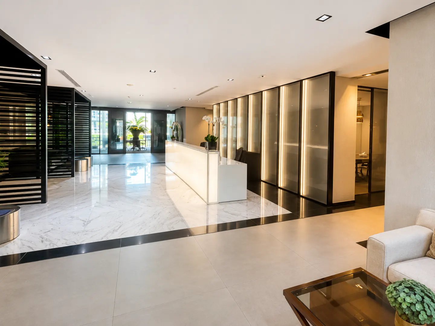 Modern office lobby with a glowing white reception desk, marble floors, black slatted partitions, and a seating area.